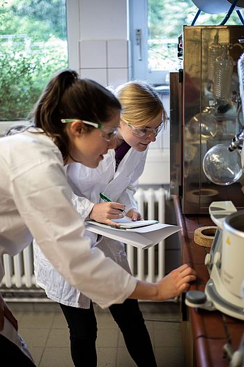 Zwei Studenten in Laborkitteln und mit Schutzbrillen betrachten ein Gerät